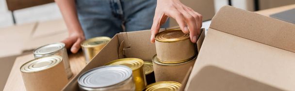 cropped view of african american volunteer packing canned food in box in charity storehouse, banner - Foto, afbeelding