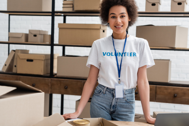 young african american volunteer with name tag smiling at camera near carton boxes - Foto, imagen