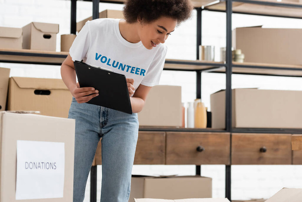 young african american woman with clipboard looking at carton boxes in donation warehouse - 写真・画像