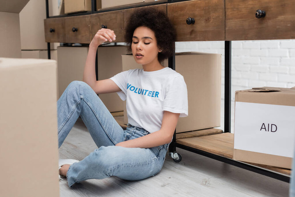 exhausted african american volunteer with closed eyes sitting on floor in charity warehouse - 写真・画像