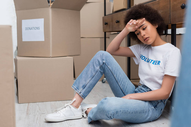 tired african american woman sitting on floor near donation boxes in charity center - Foto, afbeelding