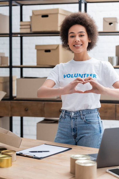 happy african american volunteer showing heart sign near clipboard on desk  - Photo, image