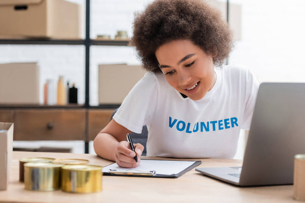smiling african american volunteer writing on clipboard near blurred laptop - Photo, image