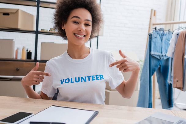 smiling african american woman pointing at t-shirt with volunteer lettering  - 写真・画像
