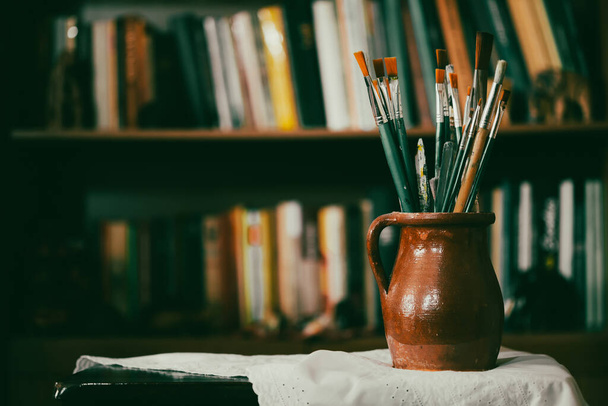 Still life of paint brushes  in clay jug, old books in the background. - Фото, зображення