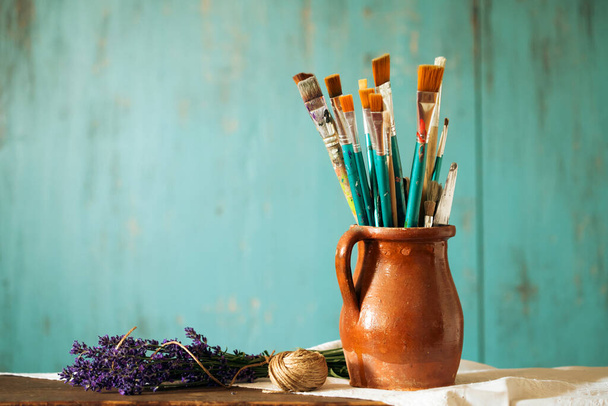 Still life of paint brushes  in clay jug an lavender flower on vintage turquoise background - Fotografie, Obrázek