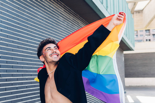Delighted gay with rhinestones on face and colorful rainbow LGBT flag in hands looking up while standing near building on street - Foto, immagini