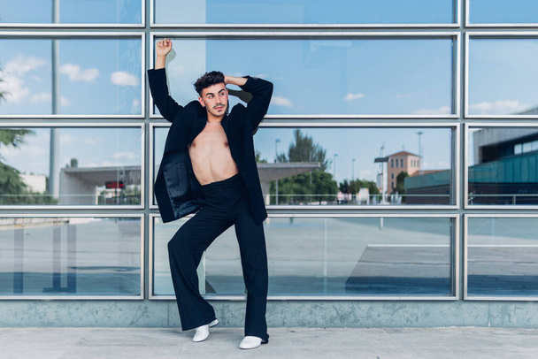 Full body of queer male in black jacket on naked torso standing with raised arm near glass wall in city - Фото, изображение