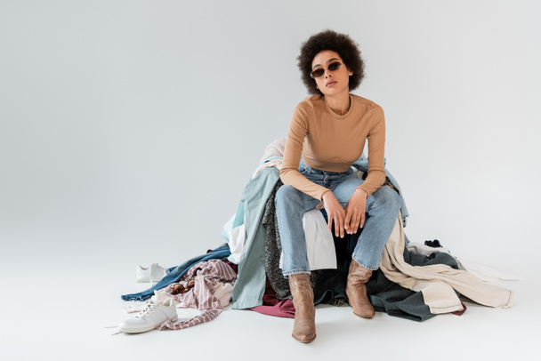 trendy african american woman in sunglasses sitting near mess of different clothes on grey background - Foto, imagen
