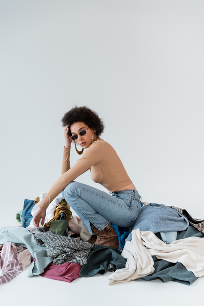 curly african american woman in sunglasses posing near assortment of clothes on grey background - Foto, imagen