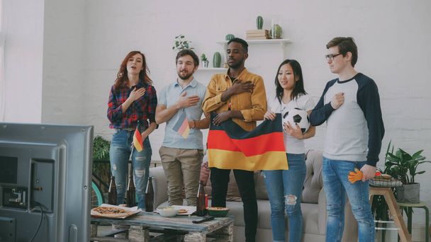 Multi-ethnic group of friends sport fans listening and singing German national anthem before watching sports championship on TV together at home indoors - Valokuva, kuva
