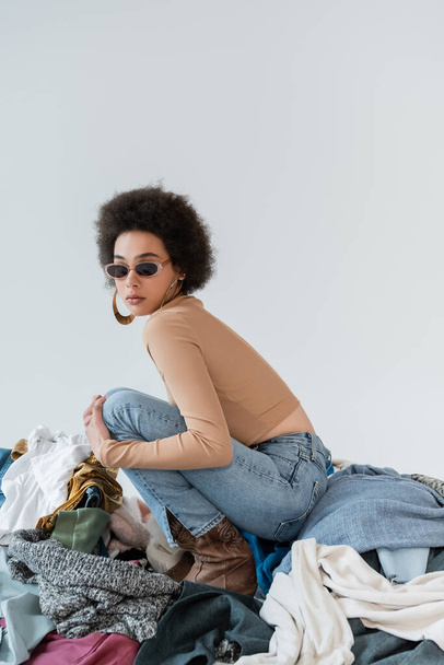 brunette african american woman in trendy sunglasses near pile of different clothes on grey background - Fotó, kép