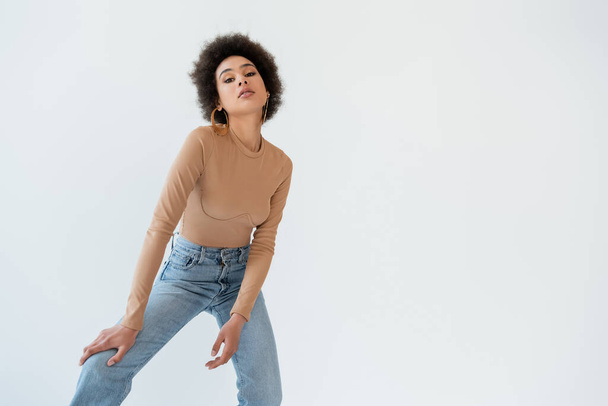 trendy african american woman in jeans and beige long sleeve shirt looking at camera isolated on grey - Fotó, kép