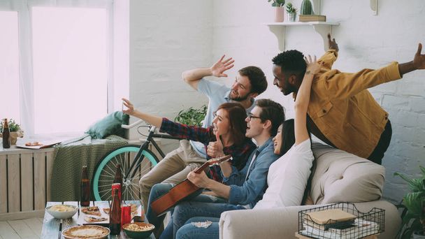 Multi ethnic group of cheerful friends taking selfie photos on smartphone camera while celebrating at party with beer and snacks at home - 写真・画像