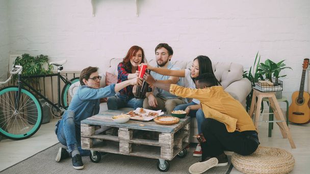 Happy friends have party talking and eating pizza. They clink bottles sitting on sofa at home indoors - 写真・画像
