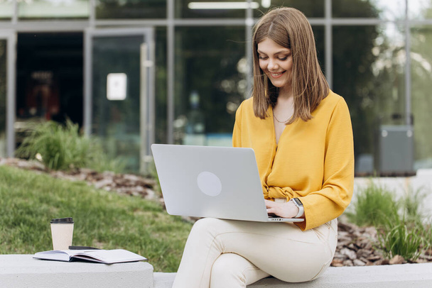 Attractive young female using laptop while sitting on bench outdoors in city. Pretty girl student preparing for classes in nature. Lady corporate start up office employee working outdoor - Valokuva, kuva
