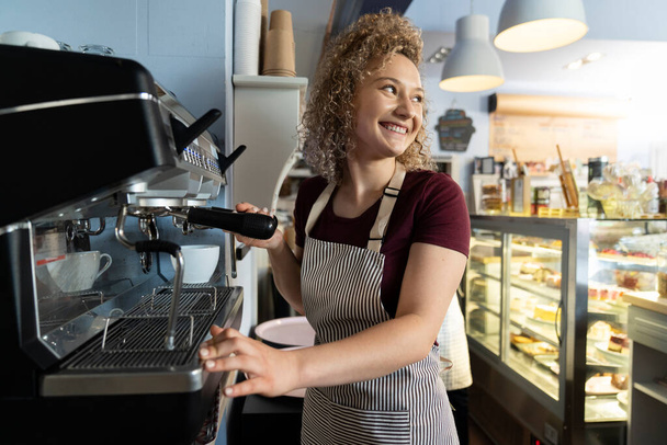 Młoda biała barmanka robi kawę w kawiarni i odwraca wzrok uśmiechem - Zdjęcie, obraz