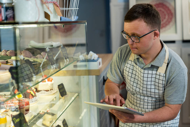 Blanke man met down syndroom doet koelkast inventarisatie in een cafe met behulp van digitale tablet - Foto, afbeelding