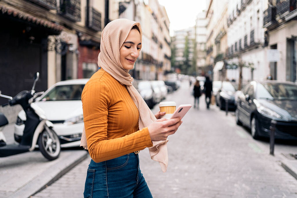Pretty young muslim woman wearing head scarf using her mobile phone and walking in the city. - Foto, imagen