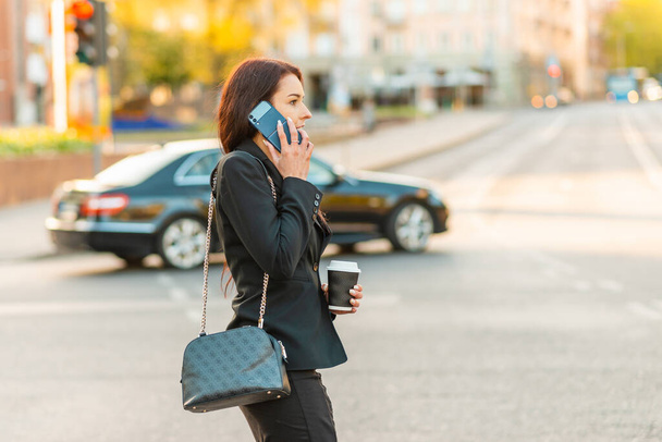 Young business woman talking on the phone holding coffee paper cup,crossing street blurred background. Fashion business photo of beautiful girl in black suite with phone and cup of coffee.Side view. - Фото, изображение