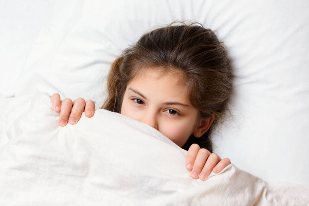 Playful little girl with charming glance lies on white pillow and looking to the camera from under the blanket, enjoying and resting on a comfortable white bed. Good morning and restful night concept - Photo, Image