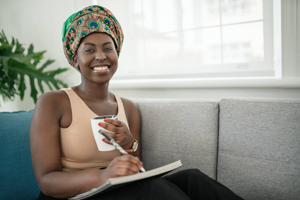 African woman studying from home, writing down notes in book. Relaxing on sofa at home. Looking into camera - Foto, afbeelding