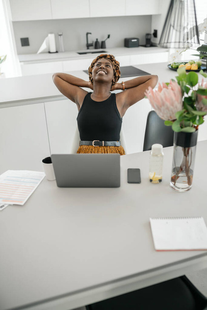 Αφρικανική παραδοσιακή επιχειρηματίας που εργάζεται από το σπίτι. Κοιτάζοντας ψηλά, τεντωμένη και τα χέρια πίσω από την ημέρα της ονειρεύεται, φορώντας εμπνευσμένο μαντήλι κεφαλής - Φωτογραφία, εικόνα