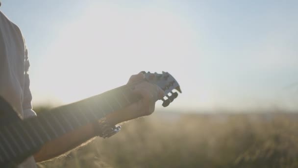 Man gitarzysta gra na gitarze w trawiastym polu o zachodzie słońca - podświetlenie strzał - Poziomy 4K wideo - Materiał filmowy, wideo