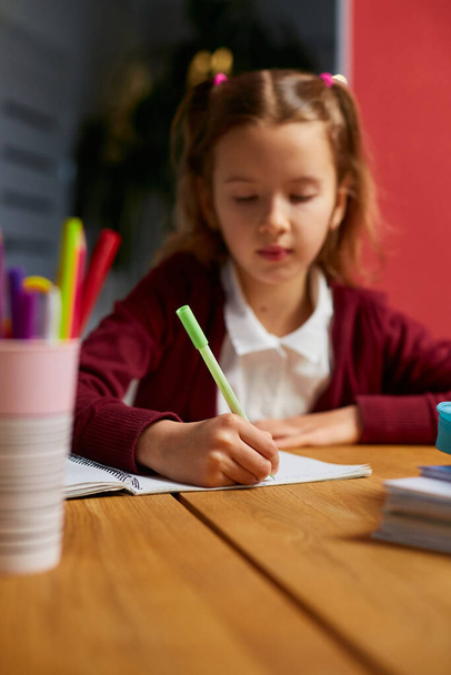 Focused schoolgirl sit at desk doing homework handwriting, homeschooling. Small junior girl engaged in distant learning get remote elementary education and preparing for exams. - Photo, Image