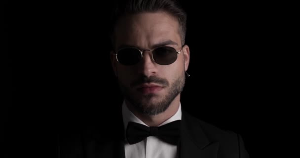 unshaved young groom wearing sunglasses, looking to side and arranging bowtie, smiling in front of black background - Imágenes, Vídeo