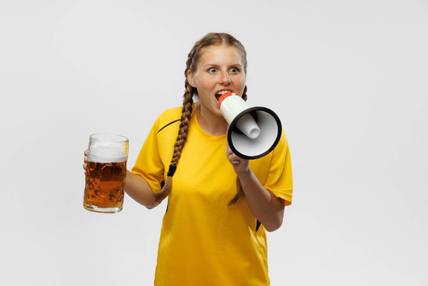 Shouting at megaphone. Young excited girl in yellow football kit holding beer mug and supports favorite team isoltaed over white background. Soccer fans, competition, sport, oktoberfest concept. - 写真・画像