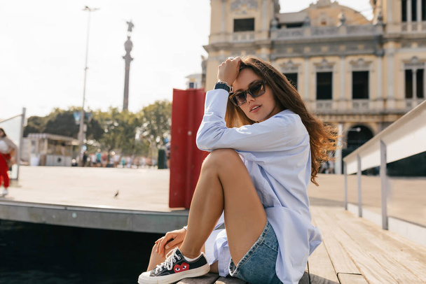 Full-length photo of happy young caucasian woman in sunglasses laughing and sitting on the pier outdoors. Brunette in good mood wears t-shirt summer. Lifestyle, different emotions, leisure concept. - Foto, afbeelding