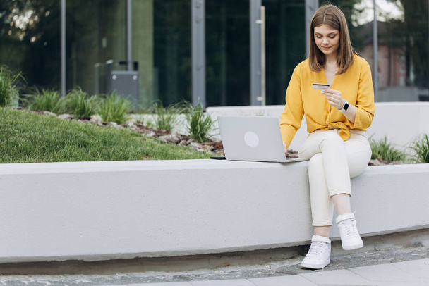 Female shopper using instant easy mobile payments making purchase in online store. E-banking app service. Girl holding credit card and smartphone sitting on bench city buildings outdoor. - Photo, image