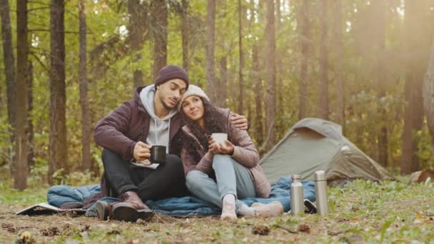 Young hispanic married couple tourists rest in woods camping man and woman in love drinking hot tea talking cute loving guy hugging girl spend nice time together smile communicate travel in nature - Metraje, vídeo