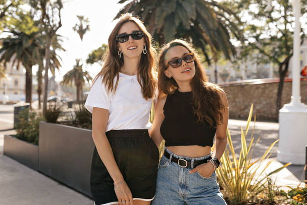 Attractive young pretty women in sunglasses wearing white and black tops posing in sunlight to camera on background of exotic plants in sunny warm day.  - Zdjęcie, obraz