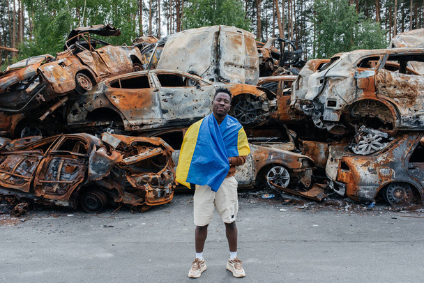 African man with Ukrainian flag stands against cars cemetery in Irpin near Kyiv out of many burnt and shelled cars. Consequences of the Russian military invasion in Ukraine. War in Ukraine and Europe. - 写真・画像
