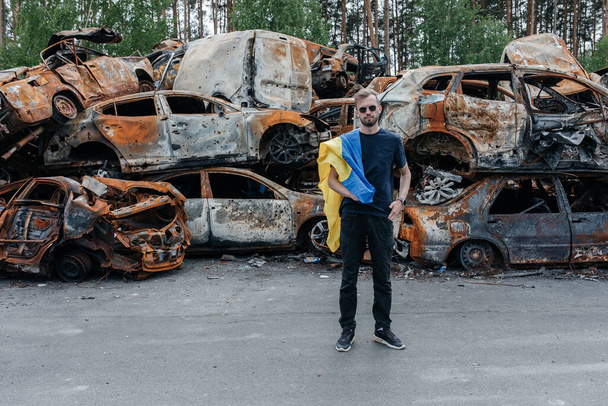 Man with Ukrainian flag stands against cars cemetery in Irpin near Kyiv out of many burnt and shelled cars. Consequences of the Russian military invasion in Ukraine. War in Ukraine and Europe. - 写真・画像
