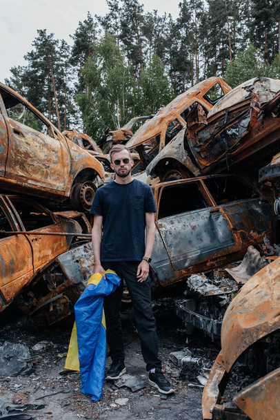 Man with Ukrainian flag stands against cars cemetery in Irpin near Kyiv out of many burnt and shelled cars. Consequences of the Russian military invasion in Ukraine. War in Ukraine and Europe. - 写真・画像