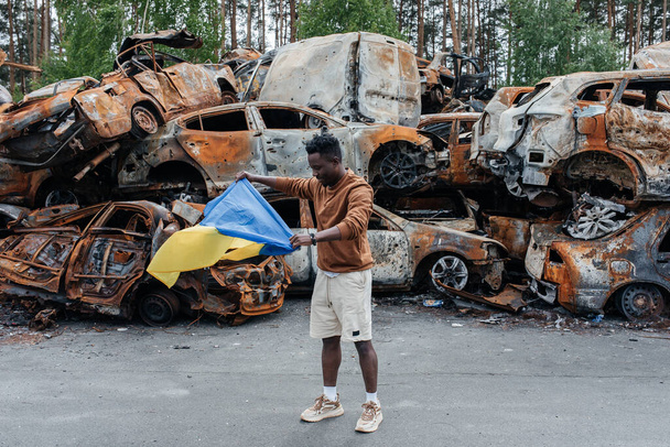African man with Ukrainian flag stands against cars cemetery in Irpin near Kyiv out of many burnt and shelled cars. Consequences of the Russian military invasion in Ukraine. War in Ukraine and Europe. - Foto, Bild