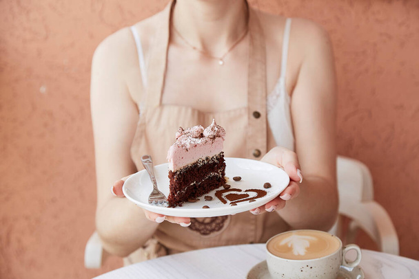Woman is holding fruit chocolate piece of cake and cup of cappuccino on wooden table outside in the cafe terrace. Atmospheric coffee time, time for yourself. Heart shaped dessert decorations - Foto, Imagem