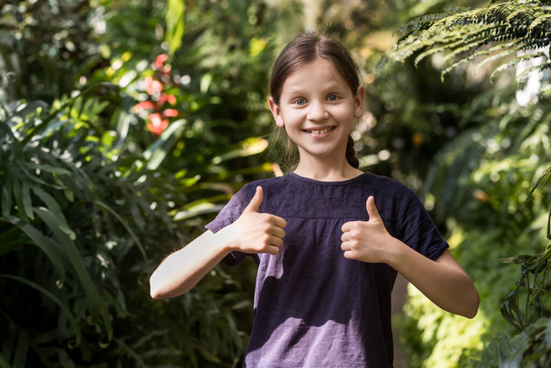 Великі пальці вгору рукою. Ботанічний сад для дітей. Щаслива дівчина з великими пальцями на фоні природи
.  - Фото, зображення