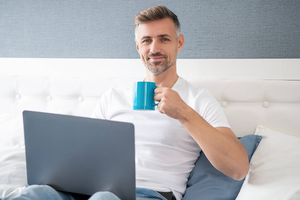positive mature man using computer in bed with coffee. - Zdjęcie, obraz