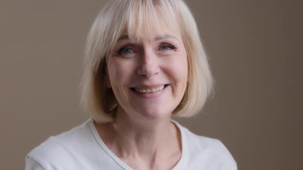 Happy mature middle-aged blonde smiling caucasian 60s woman looking at camera posing in studio portrait, cheerful older grey-haired female granny in white t-shirt, healthy carefree senior retired lady - Filmagem, Vídeo