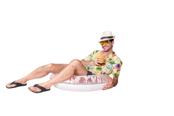 Портрет молодого улыбающегося туриста в пляжной рубашке, шляпе и солнцезащитных очках, лежащего на надувной платформе и пьющего свой кокаин. Туризм и отдых - Фото, изображение