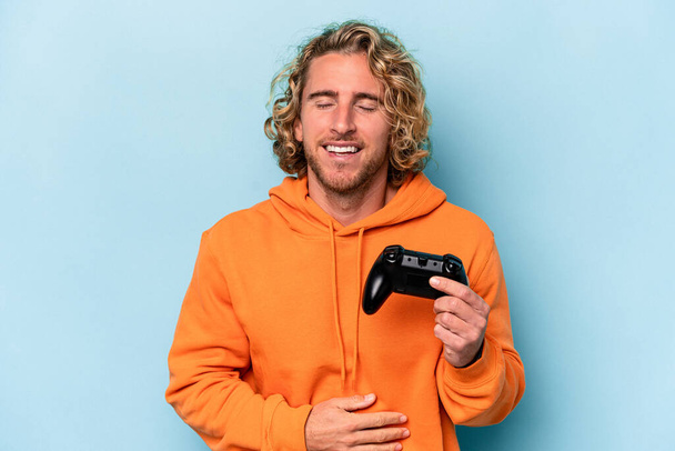 Young caucasian man playing with a video game controller isolated on blue background laughing and having fun. - Foto, Bild