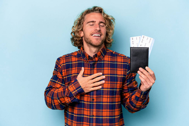Young caucasian man holding a passport isolated on blue background laughs out loudly keeping hand on chest. - Fotoğraf, Görsel