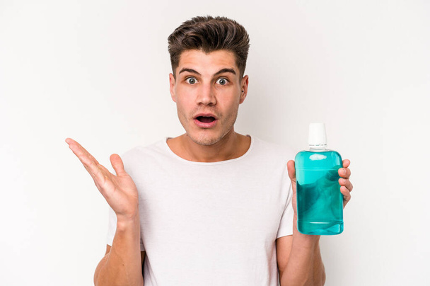 Young caucasian man holding mouthwash isolated on white background surprised and shocked. - Photo, Image
