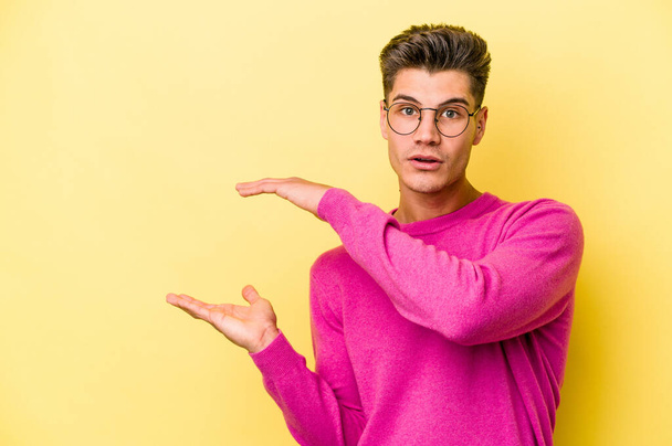 Young caucasian man isolated on yellow background shocked and amazed holding a copy space between hands. - Foto, immagini