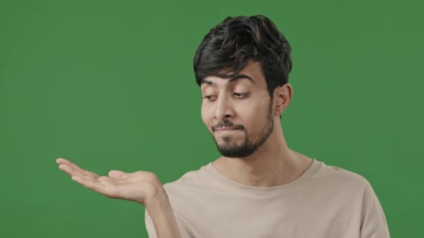 Indian young handsome man stands on green background holds copy space on hand points at empty place arabic smiling brunet guy recommend using mockup blank area for advertisement offer imaginary object - Filmagem, Vídeo