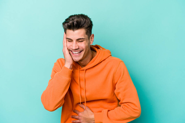 Young caucasian man isolated on blue background laughs happily and has fun keeping hands on stomach. - Photo, Image